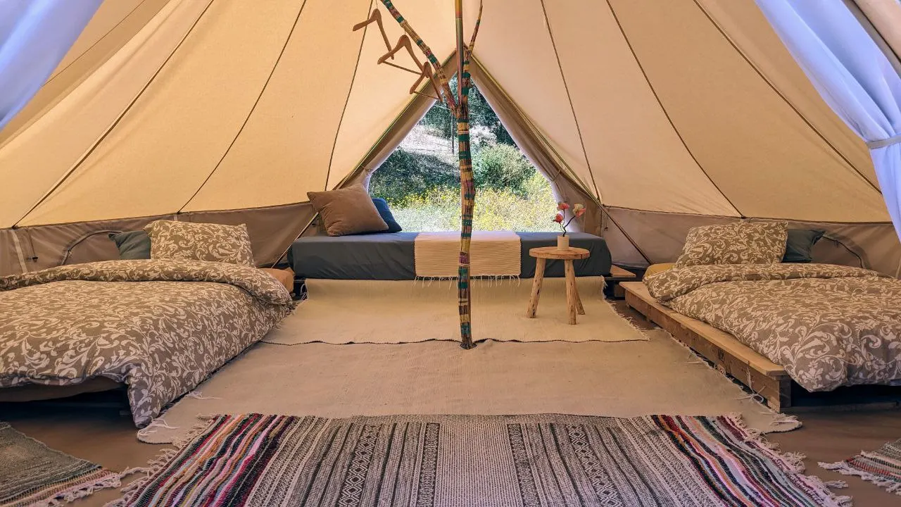 Inside a belltent: two made-up beds, woven rugs and a small table, with the doorway open to green bushes