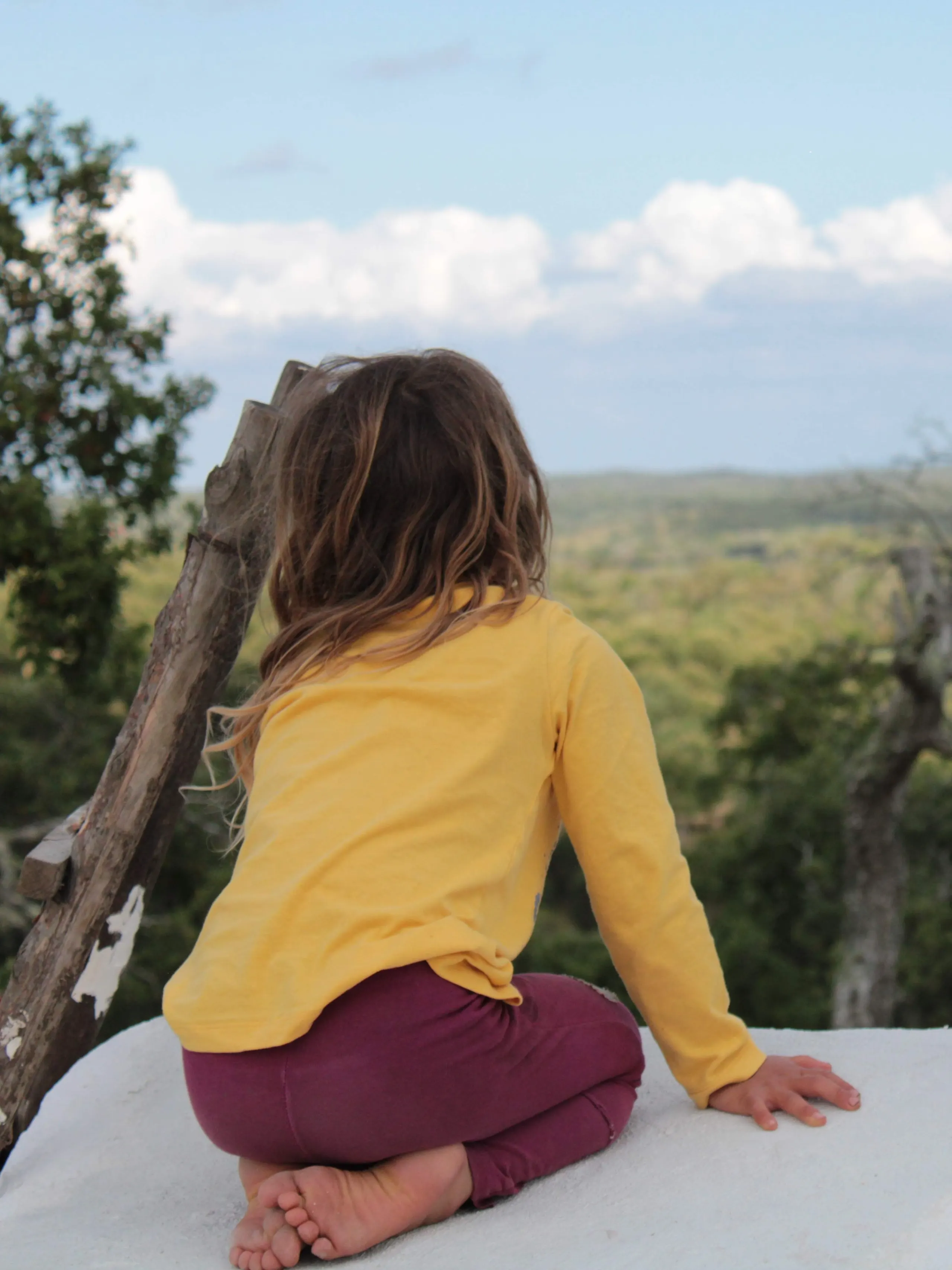A child in a yellow top sitting barefoot on a white wall, looking out over the wooded hills
