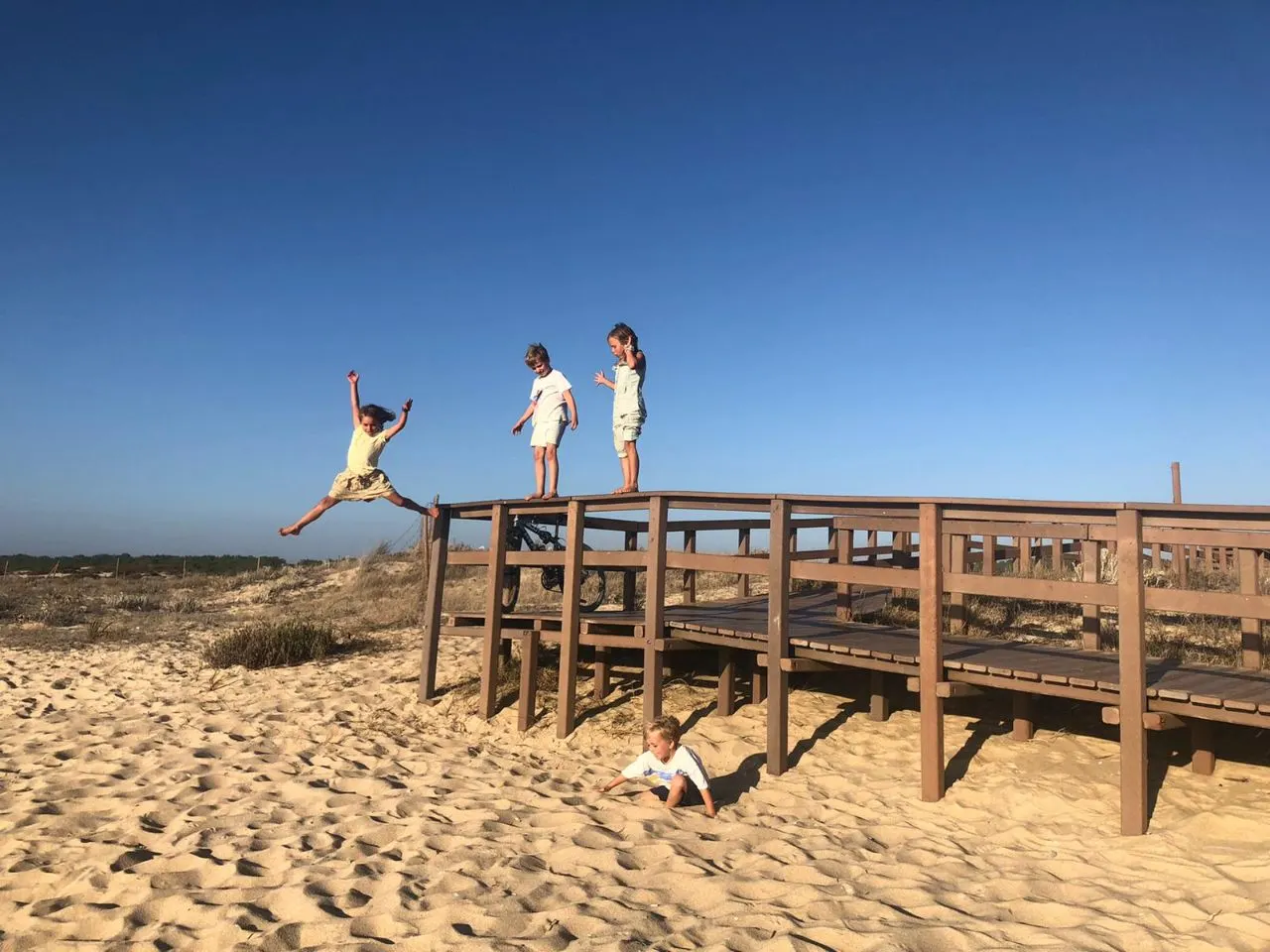 Children jumping from a wooden boardwalk into the sand dunes under a clear blue sky